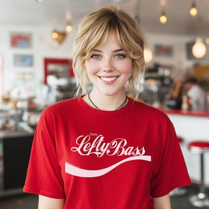 Red unisex T-shirt featuring the text "Enjoy Lefty Bass" in white and pink, worn by a smiling blonde person in a café setting.