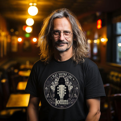 Unisex black T-shirt featuring "I Play Guitar Like Iommi" graphic, worn by a man with mid-length hair in a café setting.